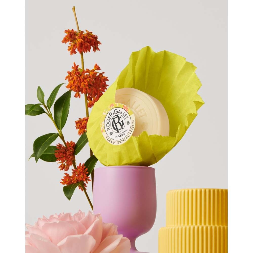 Seife in gelbem Papier, neben Blumen und einem rosa Becher. ROGER&GALLET, Fleur d'Osmanthus.