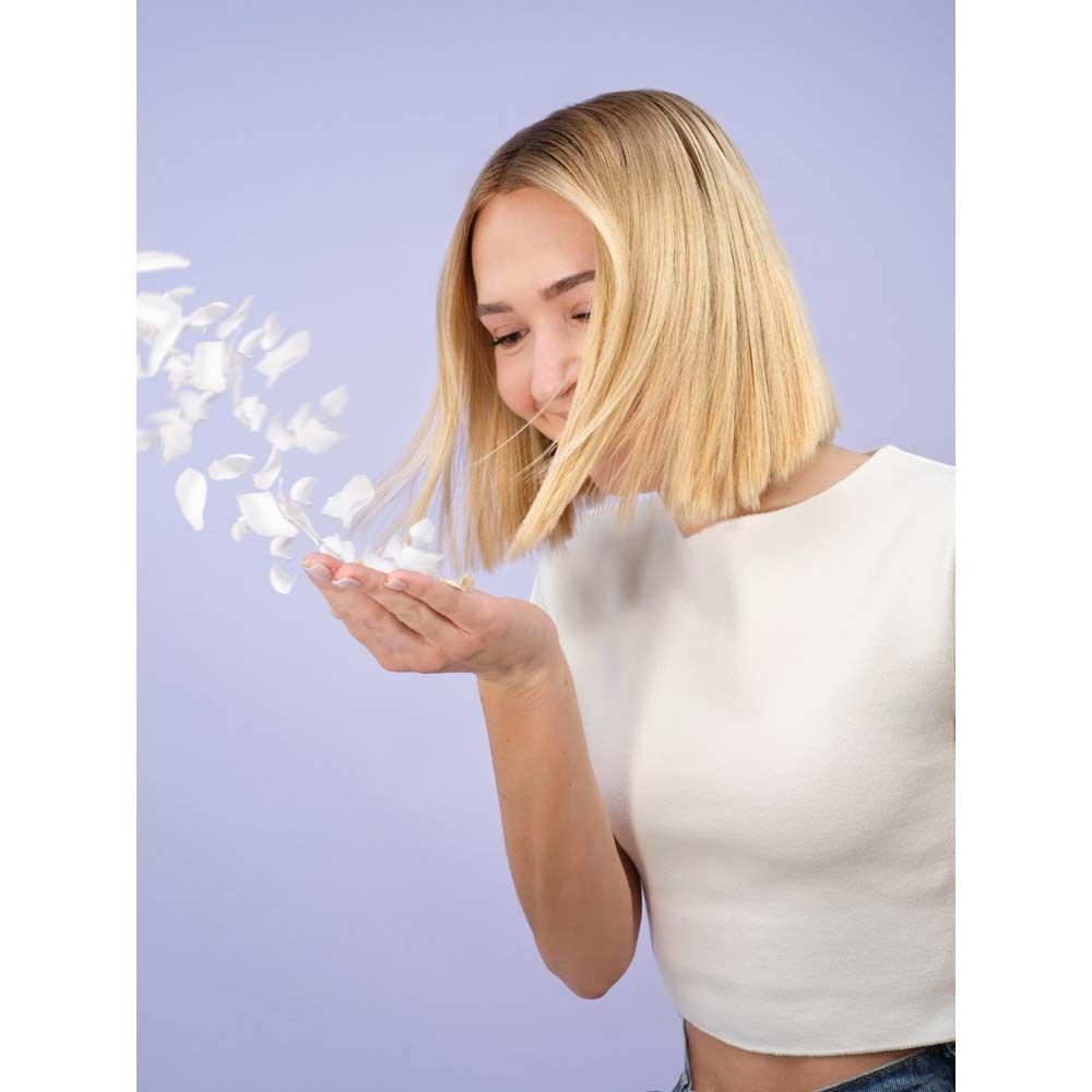 Eine Frau mit blonden Haaren hält Blütenblätter in ihrer Hand. Hintergrund ist hellviolett.