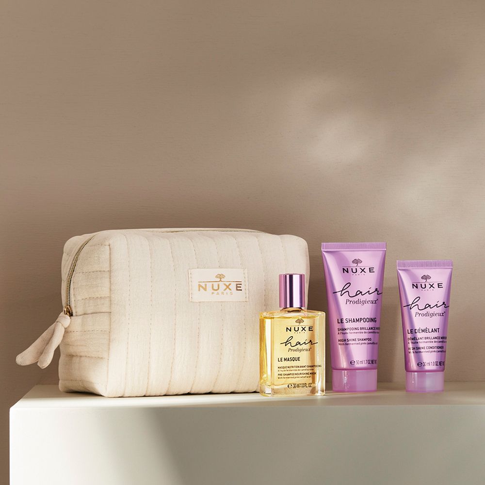 Beige Kosmetiktasche mit drei Haarprodukten. Flasche und zwei Tuben. Aufschrift: NUXE, Hair Prodigieux. Auf weißer Oberfläche.