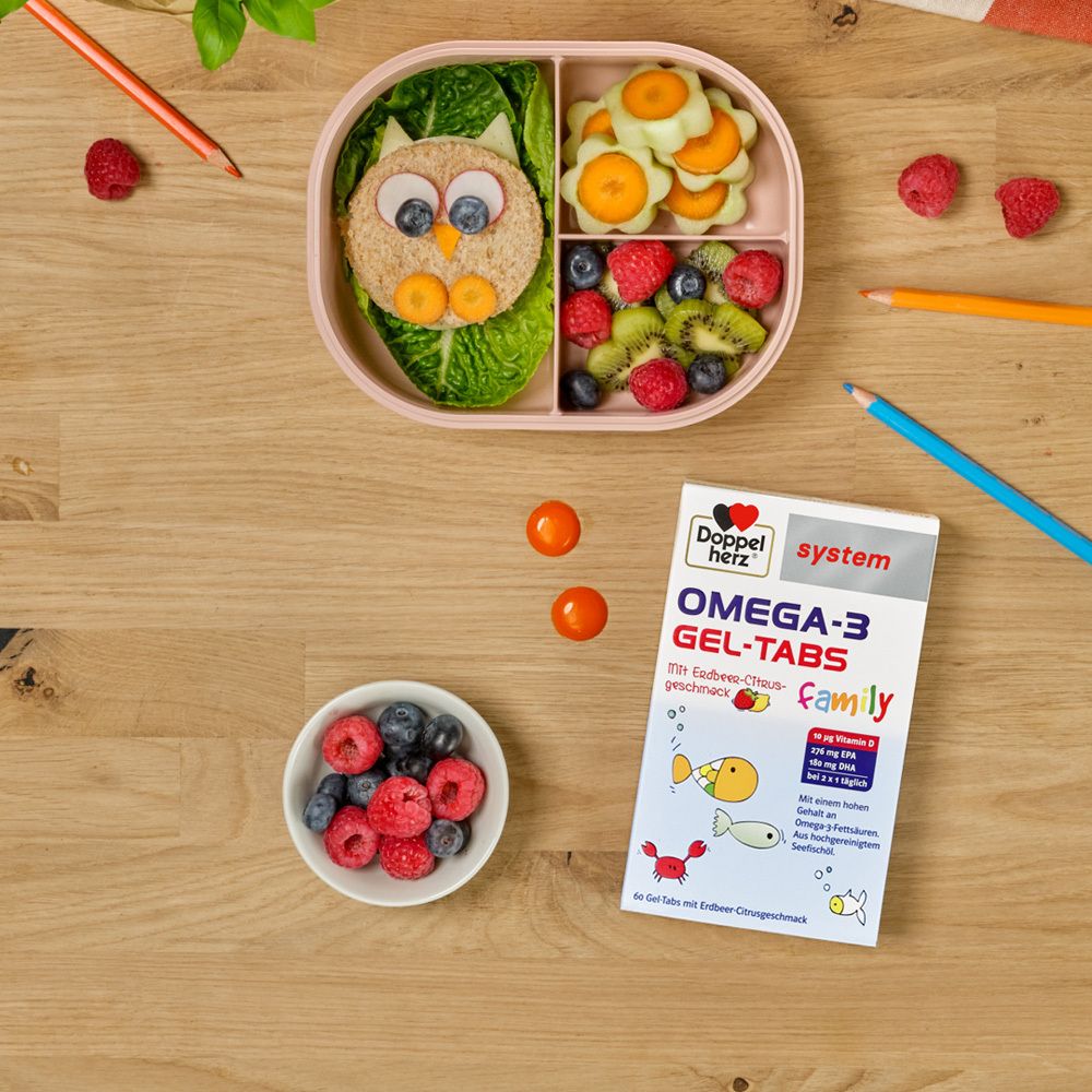 Doppelherz system OMEGA-3 GEL-TABS Family Packung neben einer Brotdose mit Essen und Obst.