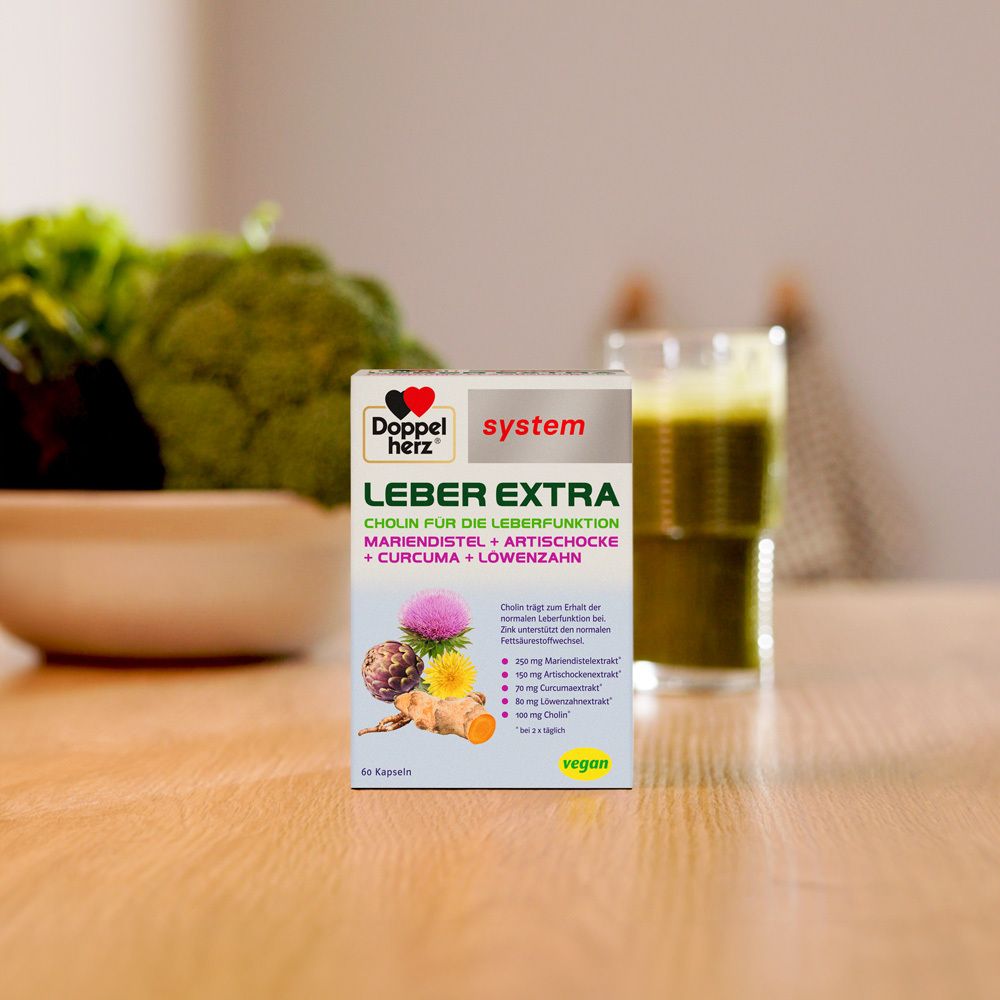 Doppelherz Leber Extra Kapseln. Packung auf Holztisch. Im Hintergrund Gemüse und ein Getränk.