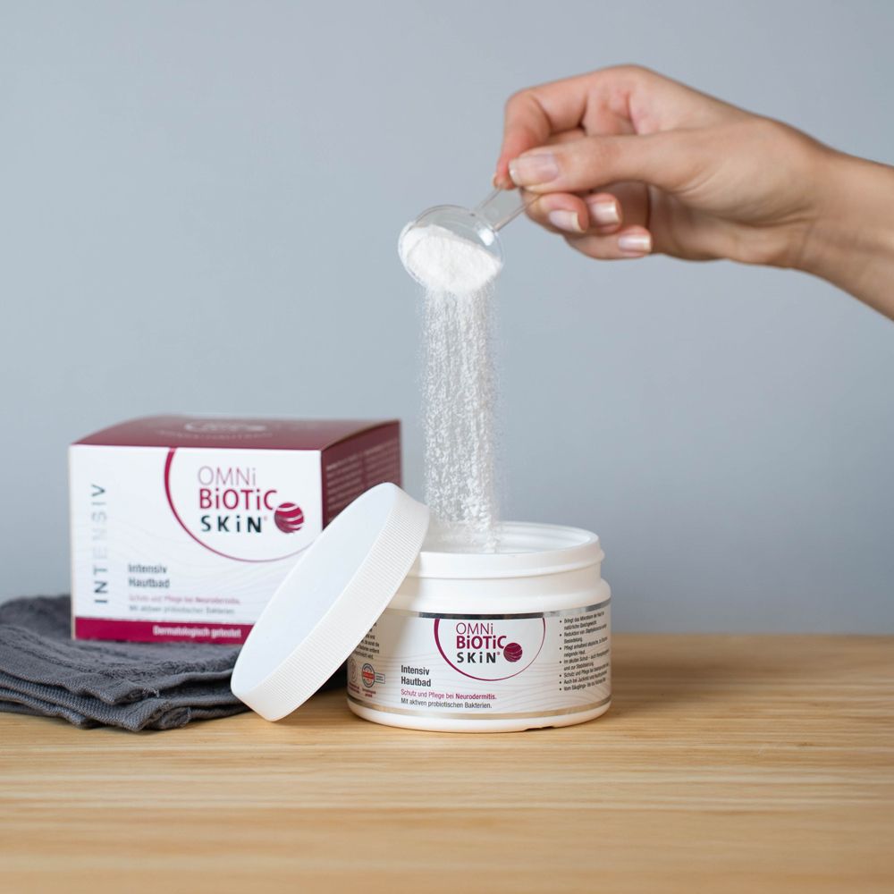 OMNi-BiOTiC SKIN® Intensiv Hautbad. Weißes Pulver wird in ein offenes Gefäß gefüllt. Daneben die Produktverpackung. Aufschrift: OMNi-BiOTiC SKIN.