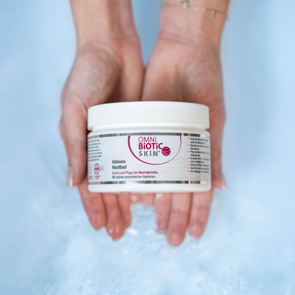 Weißes Gefäß mit OMNi-BiOTiC SKIN® Intensiv Hautbad, gehalten in Händen. Aufschrift: OMNi-BiOTiC SKIN. Hintergrund: blaues Wasser.