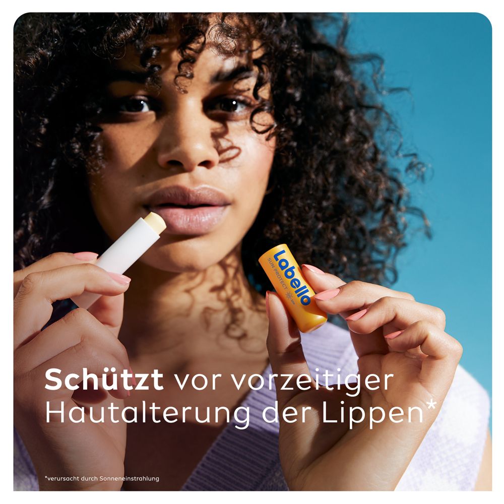 Junge Frau mit dunklen Locken trägt Labello SUN PROTECT auf die Lippen auf, hält den Lippenpflegestift.