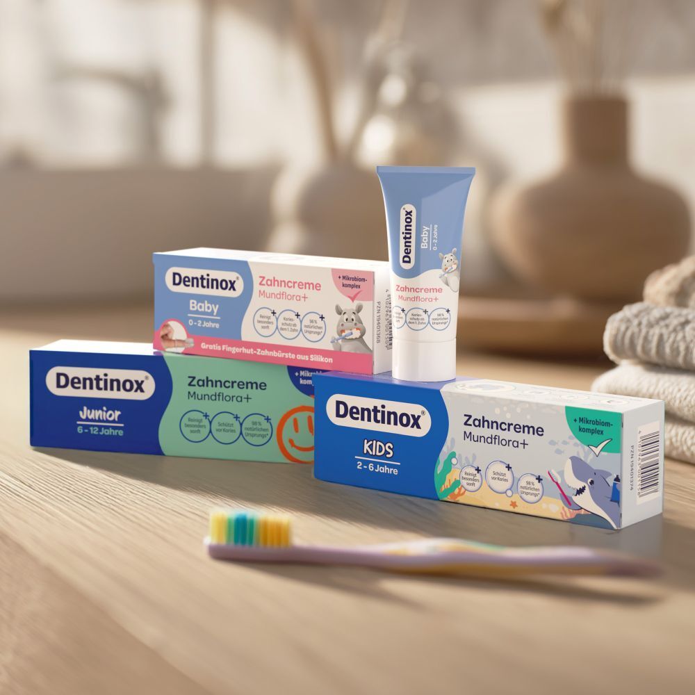 Dentinox Zahnpasta-Packungen für Baby, Junior und Kids. Eine Zahnbürste liegt daneben. Produktnamen und Illustrationen sind sichtbar.