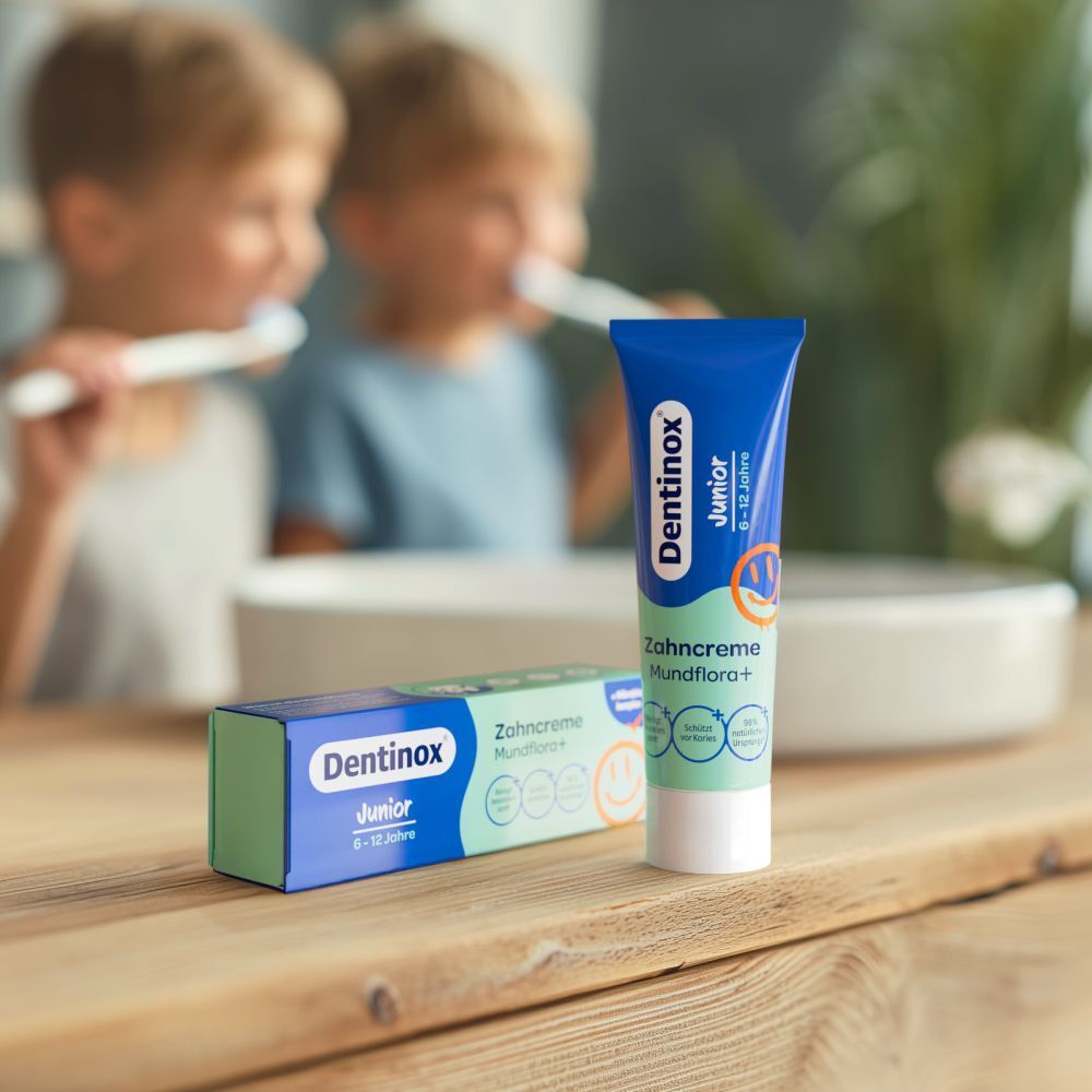 Zwei Kinder putzen Zähne. Daneben Tube und Schachtel. Aufschrift: Dentinox Junior, Zahncreme Mundflora+, 6-12 Jahre.