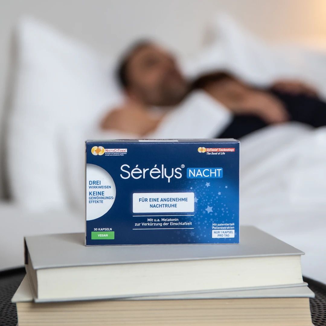 Sérélys NACHT-Verpackung auf Büchern, im Hintergrund eine Person im Bett. Blaue Verpackung mit weißen Schriftzügen.