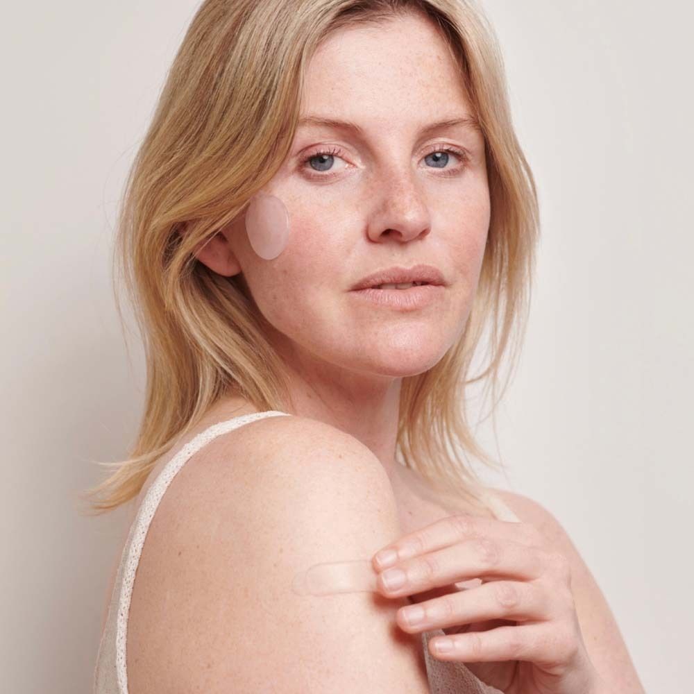 Frau mit Narben Pad auf der Wange und einem länglichen Pad am Arm. Helle Haut, blondes Haar.
