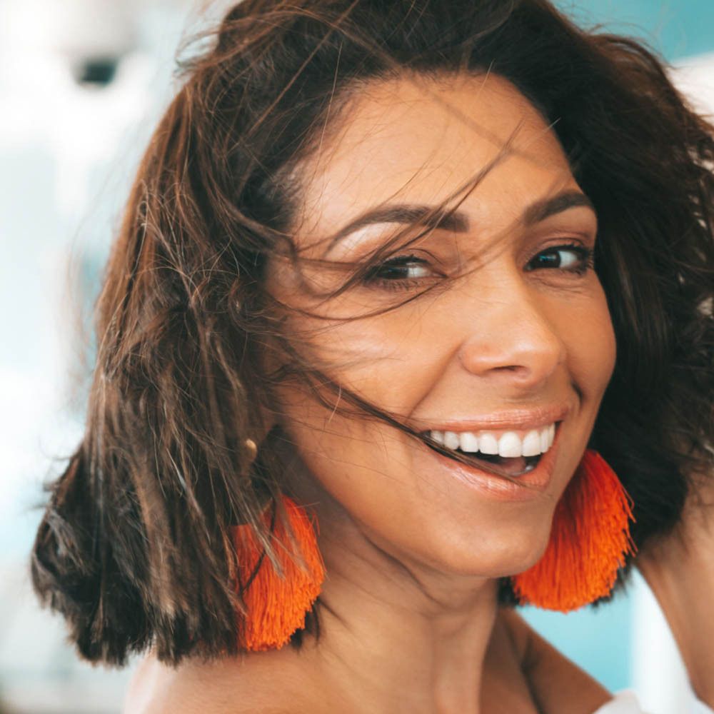 Portret van een lachende vrouw met bruin haar en oranje oorbellen.