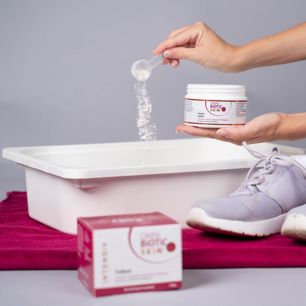 Person hält einen Messlöffel über einem Fußbad. Dose und Schachtel mit OMNi-BiOTiC SKiN® Intensiv-Fußbad.