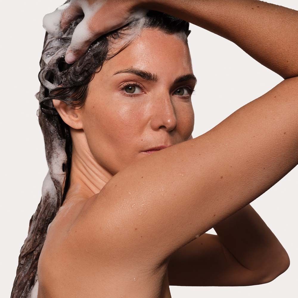 Frau wäscht sich die Haare mit Schaum. Sie schaut in die Kamera. Weißer Hintergrund.