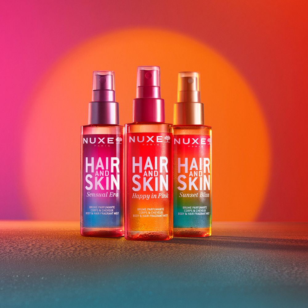 Drei Flaschen mit Sprühköpfen. Aufschrift: NUXE, HAIR AND SKIN. Verschiedene Düfte. Hintergrund: Rot, Orange, Blau.