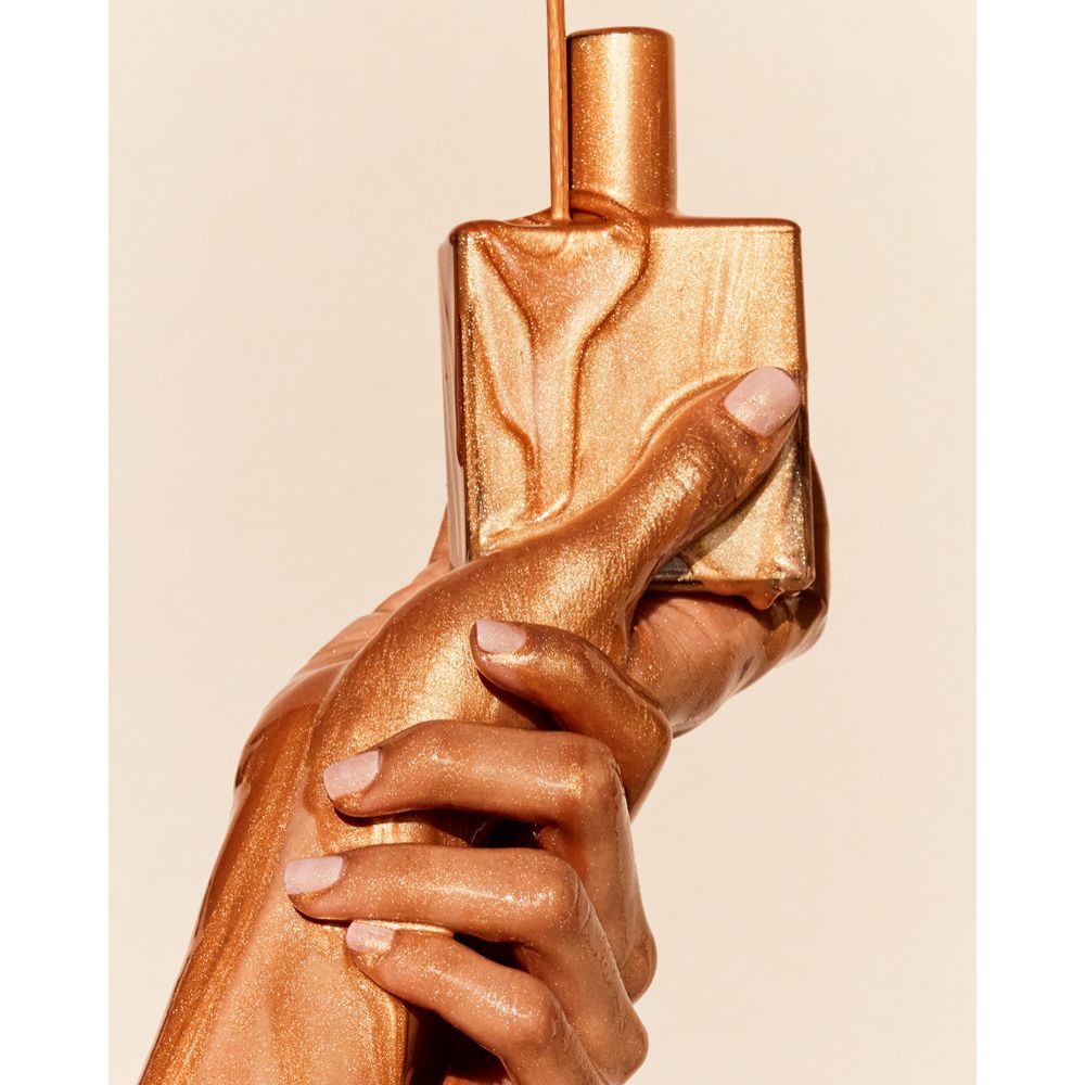 Hände halten eine goldfarbene Flasche, bedeckt mit goldfarbener Flüssigkeit. Nägel sind lackiert.