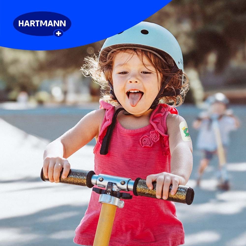 Kind auf einem Roller mit Pflaster. Pflaster mit Peppa Pig-Motiv auf dem Arm. Kind lächelt.