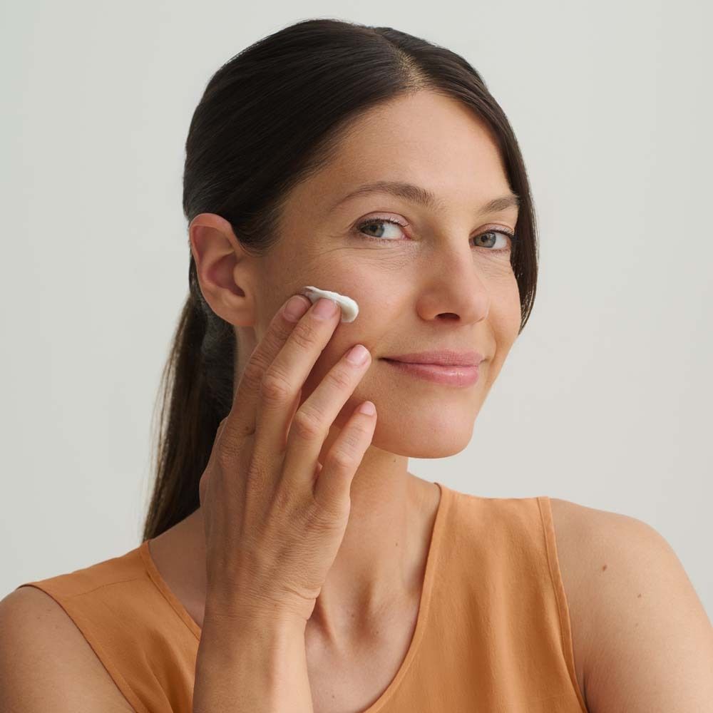 Frau trägt Creme auf ihr Gesicht auf. Sie lächelt. Hautpflege. Helle Haut. Orangefarbenes Oberteil. Hintergrund ist hell.