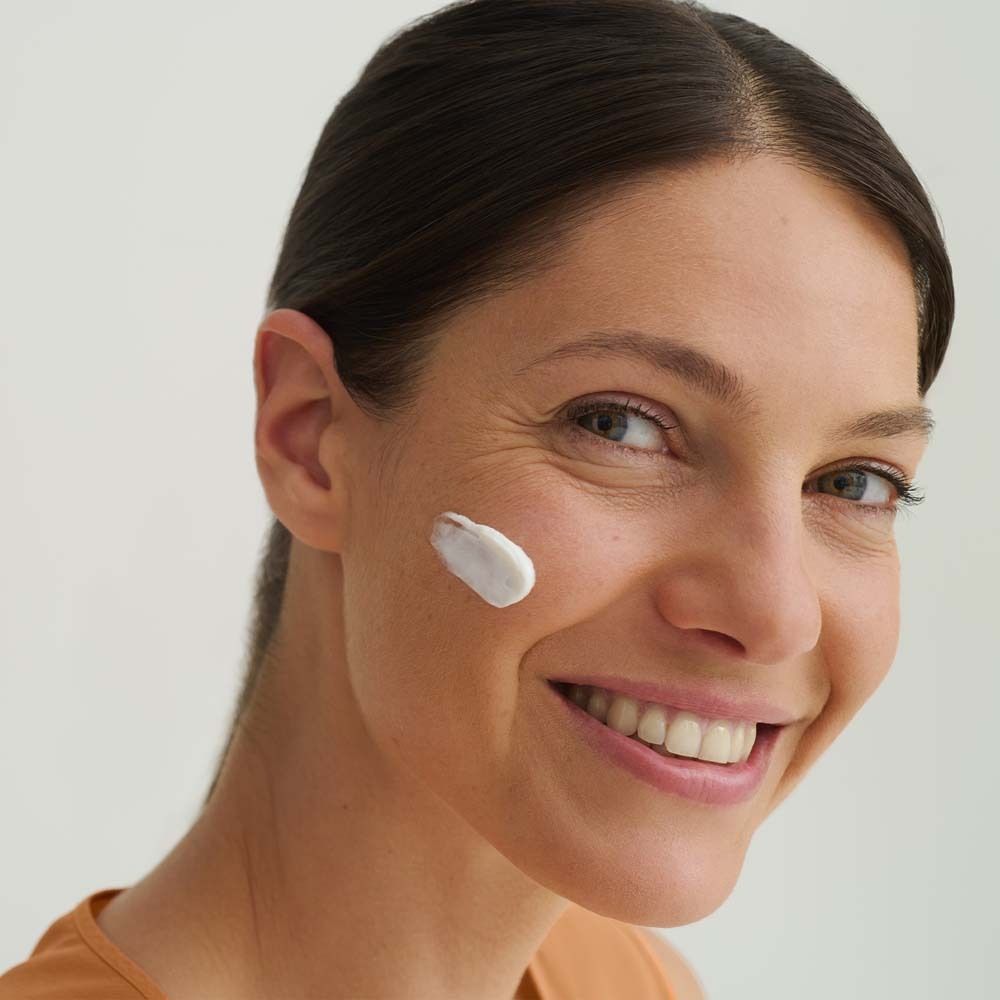 Frau mit Creme auf der Wange. Sie lächelt. Hautpflege. Helle Haut. Orangefarbenes Oberteil. Hintergrund ist hell.