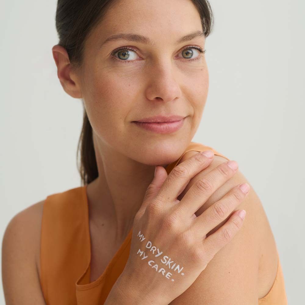Frau mit Text auf der Hand: MY DRY SKIN. MY CARE. Sie trägt ein orangefarbenes Oberteil. Hintergrund ist hell.