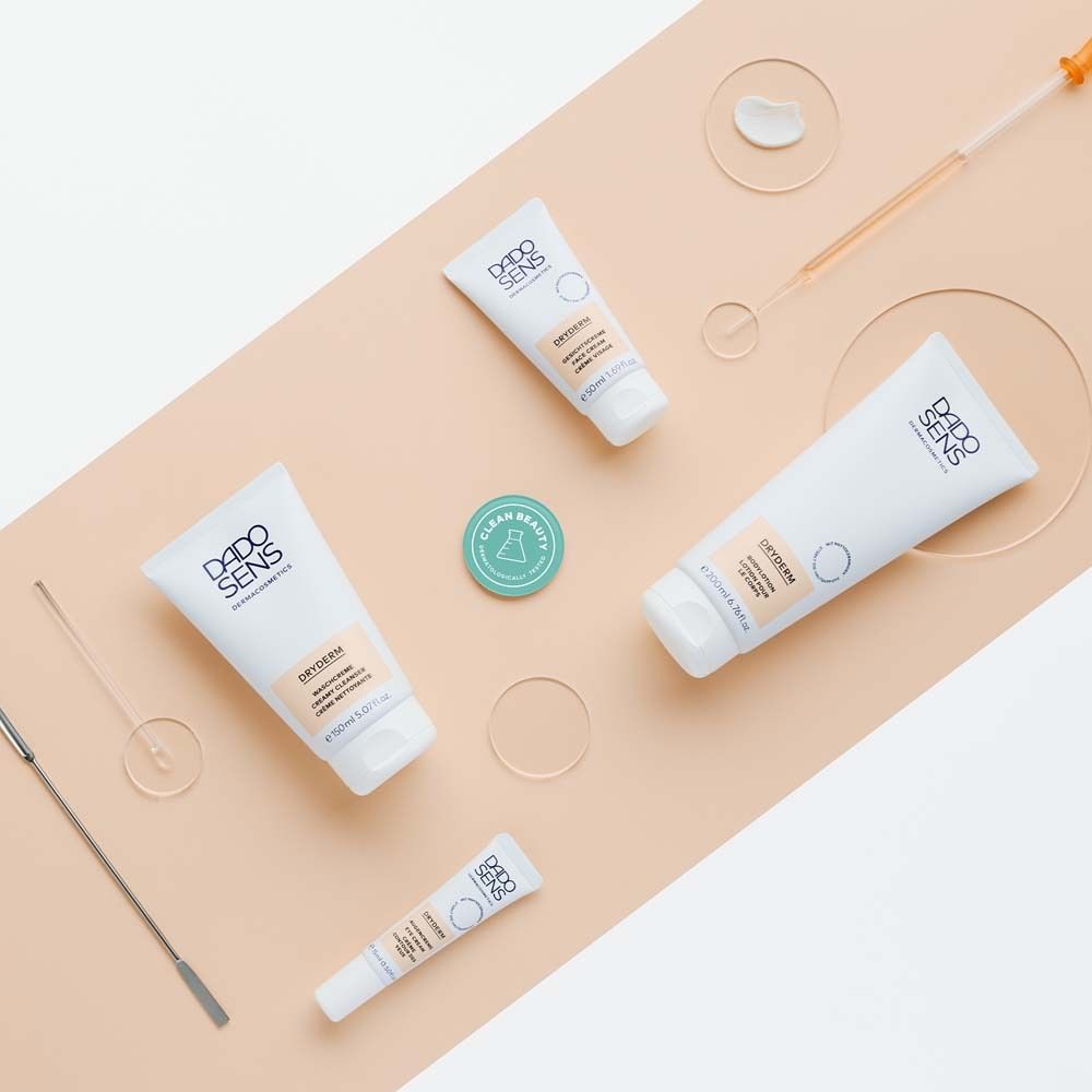 Verschiedene DADOSENS DRYDERM Produkte und Zubehör auf schrägem, beigen Hintergrund. Flaschen, Tiegel, Pipette, Spatel.