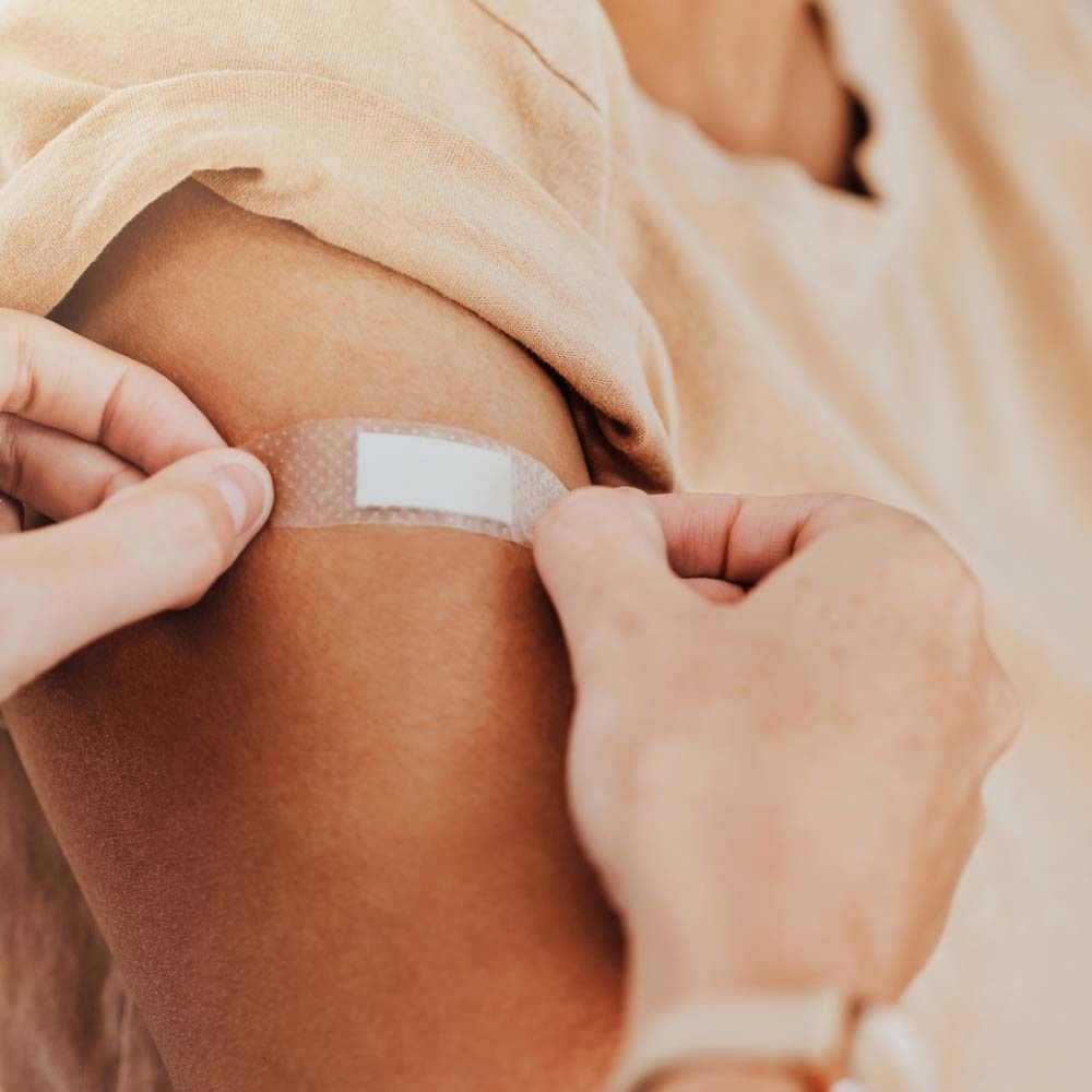 Person klebt transparentes Pflaster auf Arm. Pflaster rechteckig, mit weißem Mittelteil. Hände halten Pflaster.