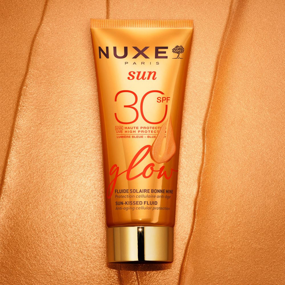 NUXE Sun Sonnenfluid Glow LSF 30. Tube auf goldenem Stoff. Text: SPF 30, glow, Fluide Solaire Bonne Mine.