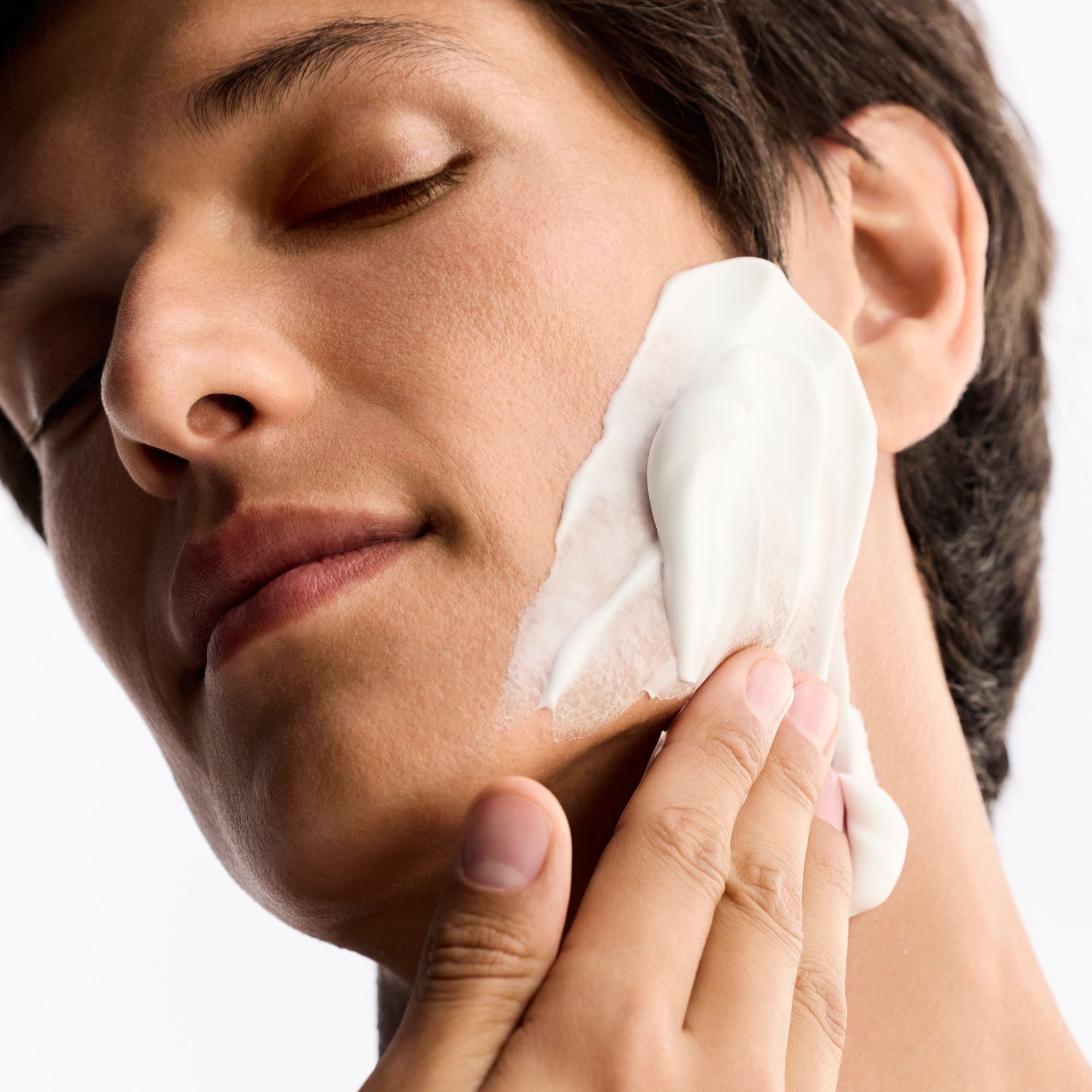 Mann mit geschlossenen Augen, der Rasiercreme auf das Gesicht aufträgt. Weiße Creme auf der Haut.