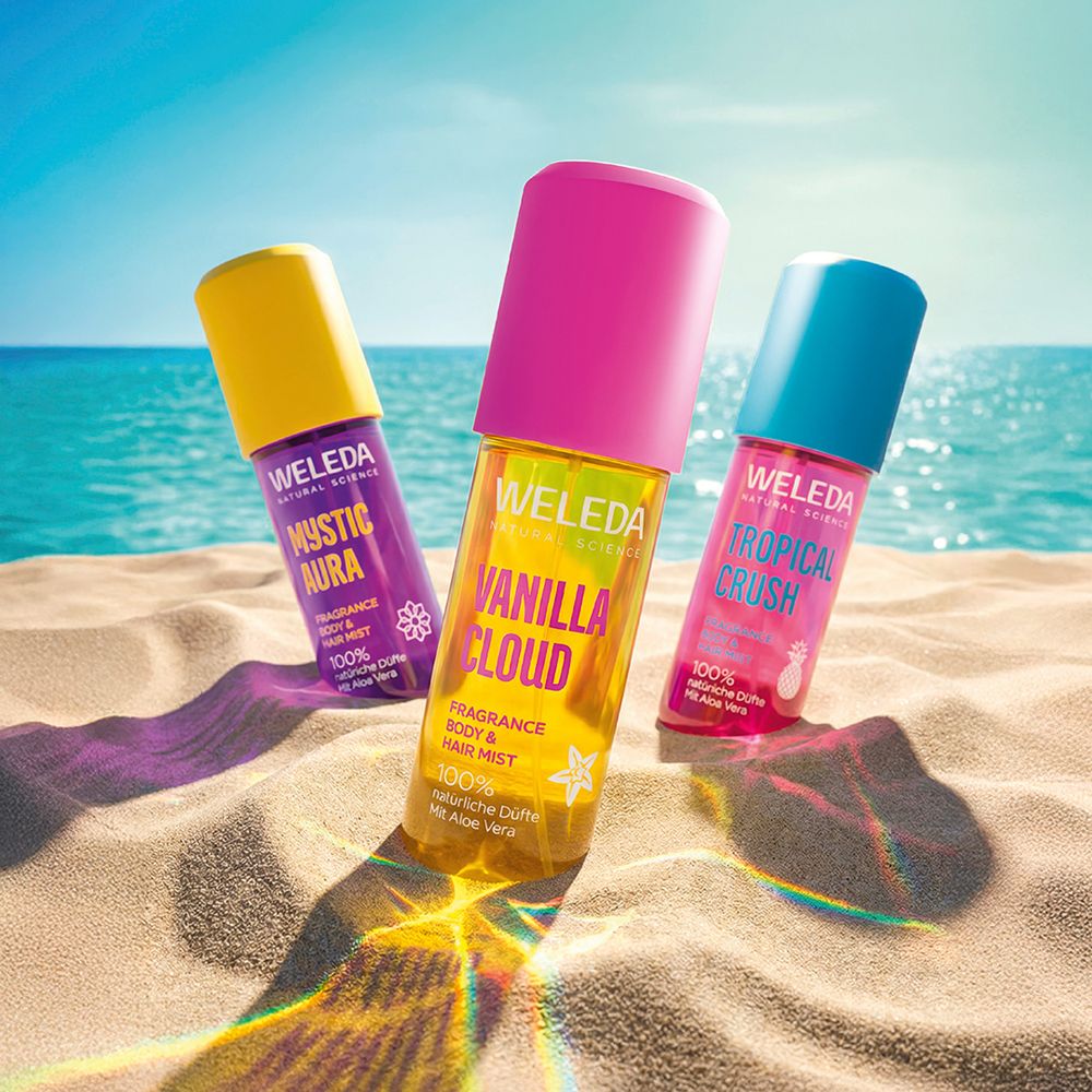 Drei Flaschen am Strand. Lila, pink und blau. Text: WELEDA, MYSTIC AURA, VANILLA CLOUD, TROPICAL CRUSH.