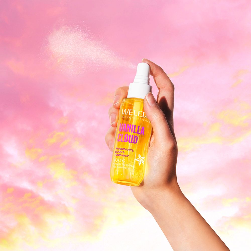 Hand sprüht Weleda Vanilla Cloud Body & Hair Mist. Hintergrund: Himmel mit Wolken.