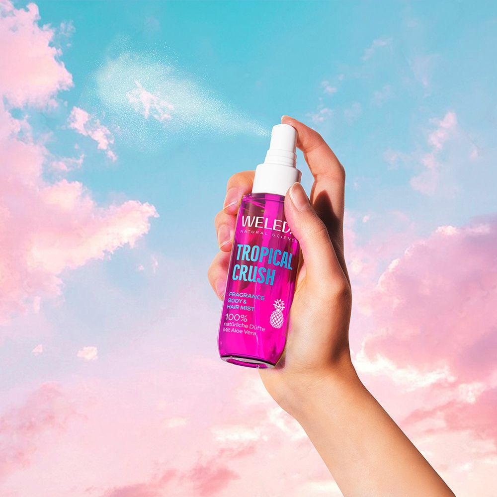 Hand sprüht Weleda Tropical Crush. Himmel Hintergrund. Pinke Flasche mit blauem Deckel.