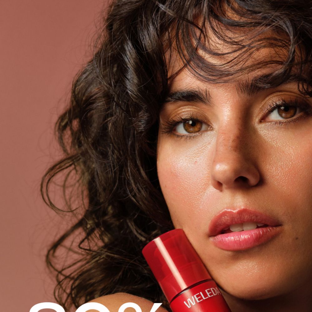 Nahaufnahme einer Frau mit dunklen Haaren, die eine rote Weleda Exo Boost Elixir Flasche hält.