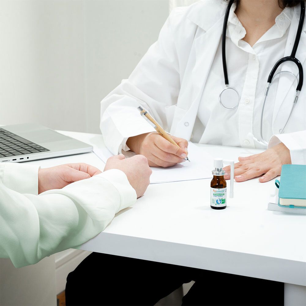 Médecin et patient à une table. Flacon avec vaporisateur blanc. PURESSENTIEL RESPIRATOIRE SPRAY GORGE. Sur la table.