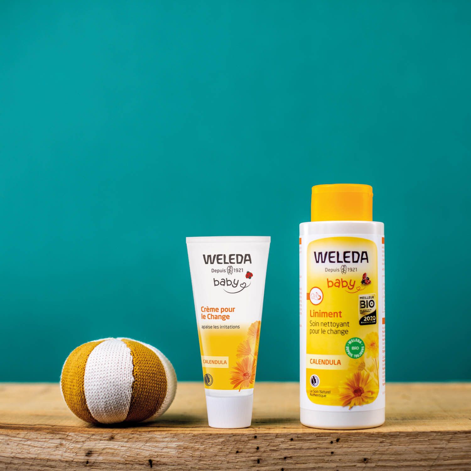 Weleda Baby-Produkte auf Holzoberfläche. Babycreme-Tube, Waschgel und ein Spielzeugball. Blauer Hintergrund.