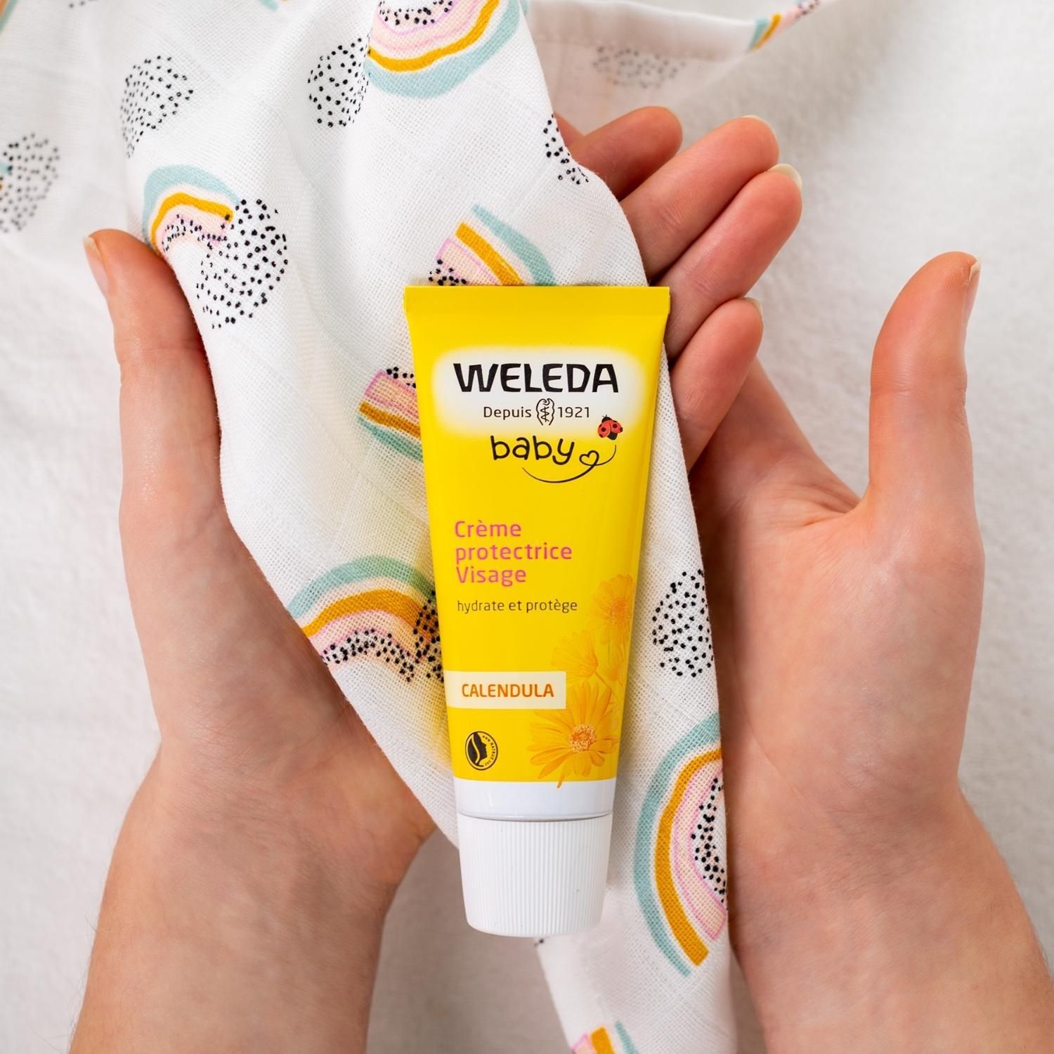 Tube de crème Weleda Baby dans les mains, entouré de tissu. Inscription: Crème protectrice Visage, Calendula.