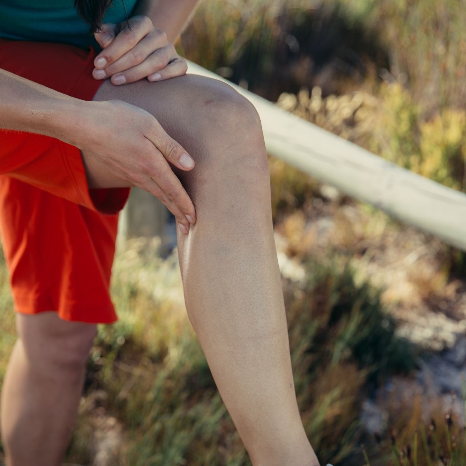 Person berührt Bein mit blauen Flecken. Person trägt rote Shorts. Natürliche Umgebung im Hintergrund.