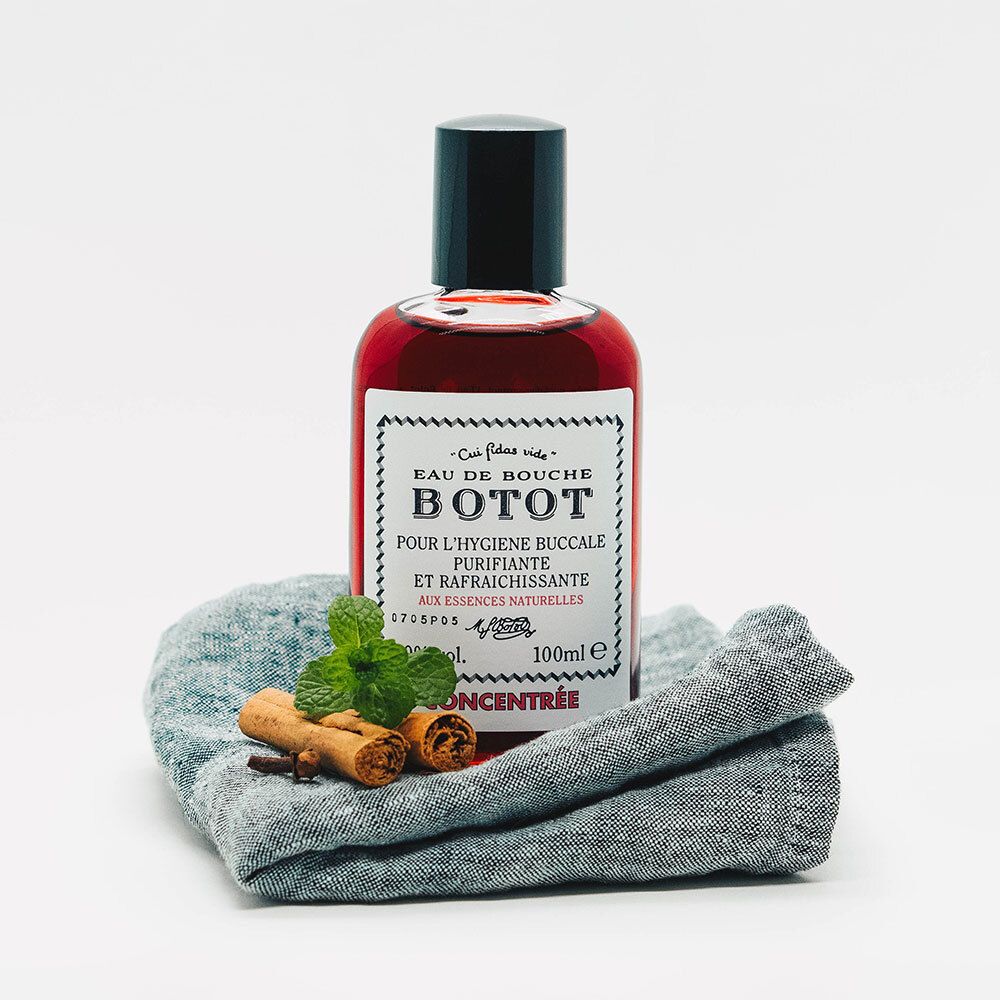 Flacon avec liquide, bouchon noir, étiquette "Botot Eau de Bouche". Sur tissu gris avec cannelle et menthe.