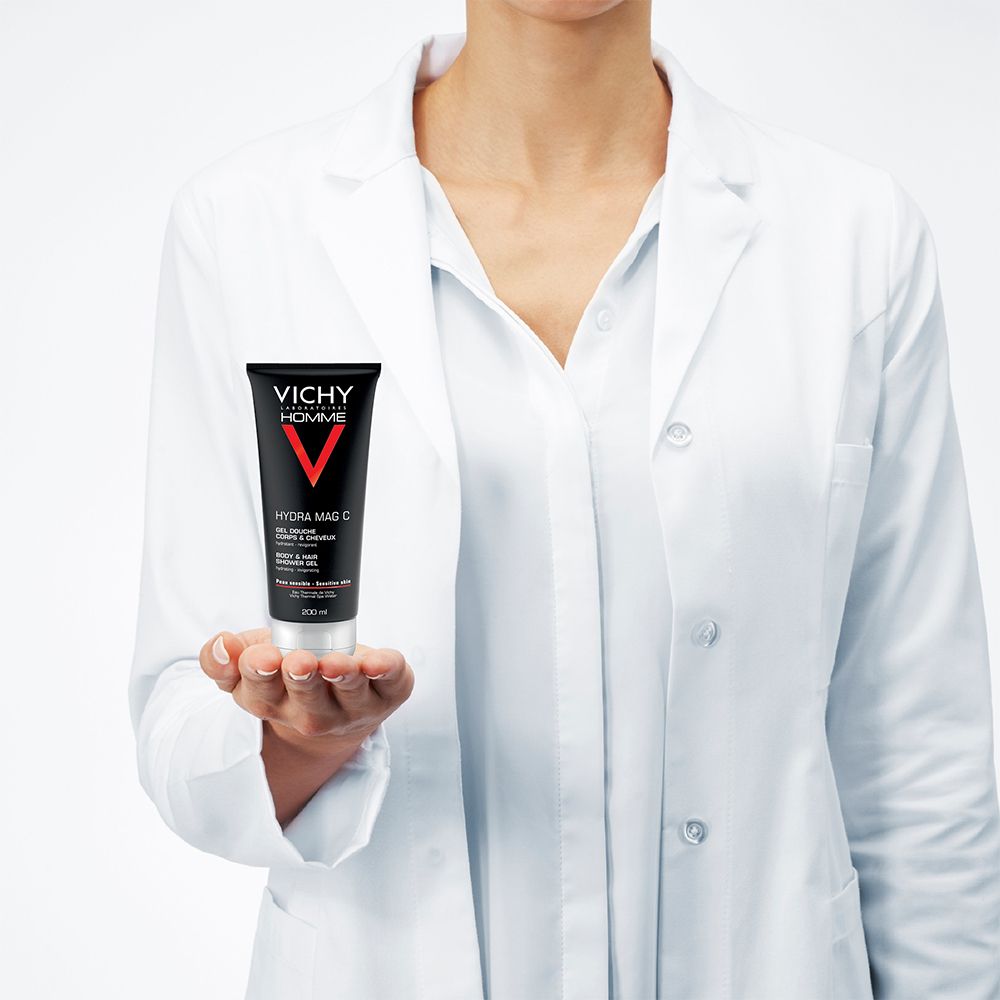 Une personne en blouse blanche tient un tube de gel douche. Tube noir avec V rouge et texte blanc. "Vichy Homme".
