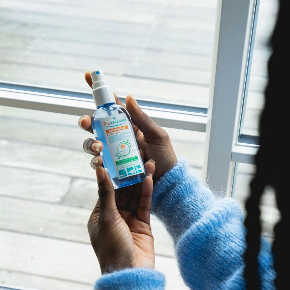 Personne tenant un spray Puressentiel Assainissant. Environnement clair avec fenêtre en arrière-plan. Vêtements bleus.