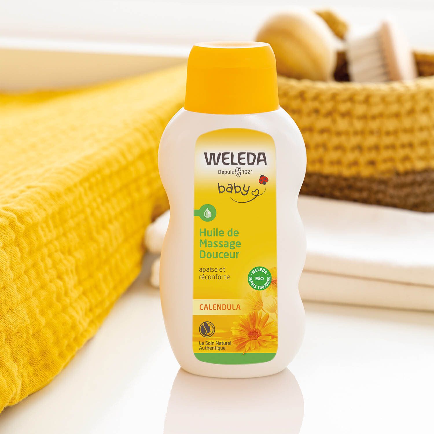 Weleda Baby Calendula Ölflasche neben gelbem Handtuch und Korb. Aufschrift: Huile de Massage Douceur, Calendula.