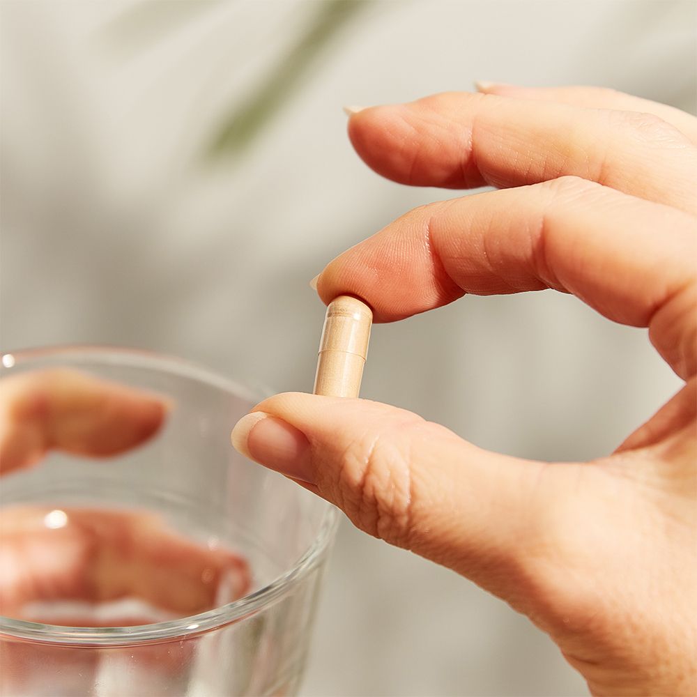 Main tenant une gélule au-dessus d'un verre d'eau. Gélule beige.