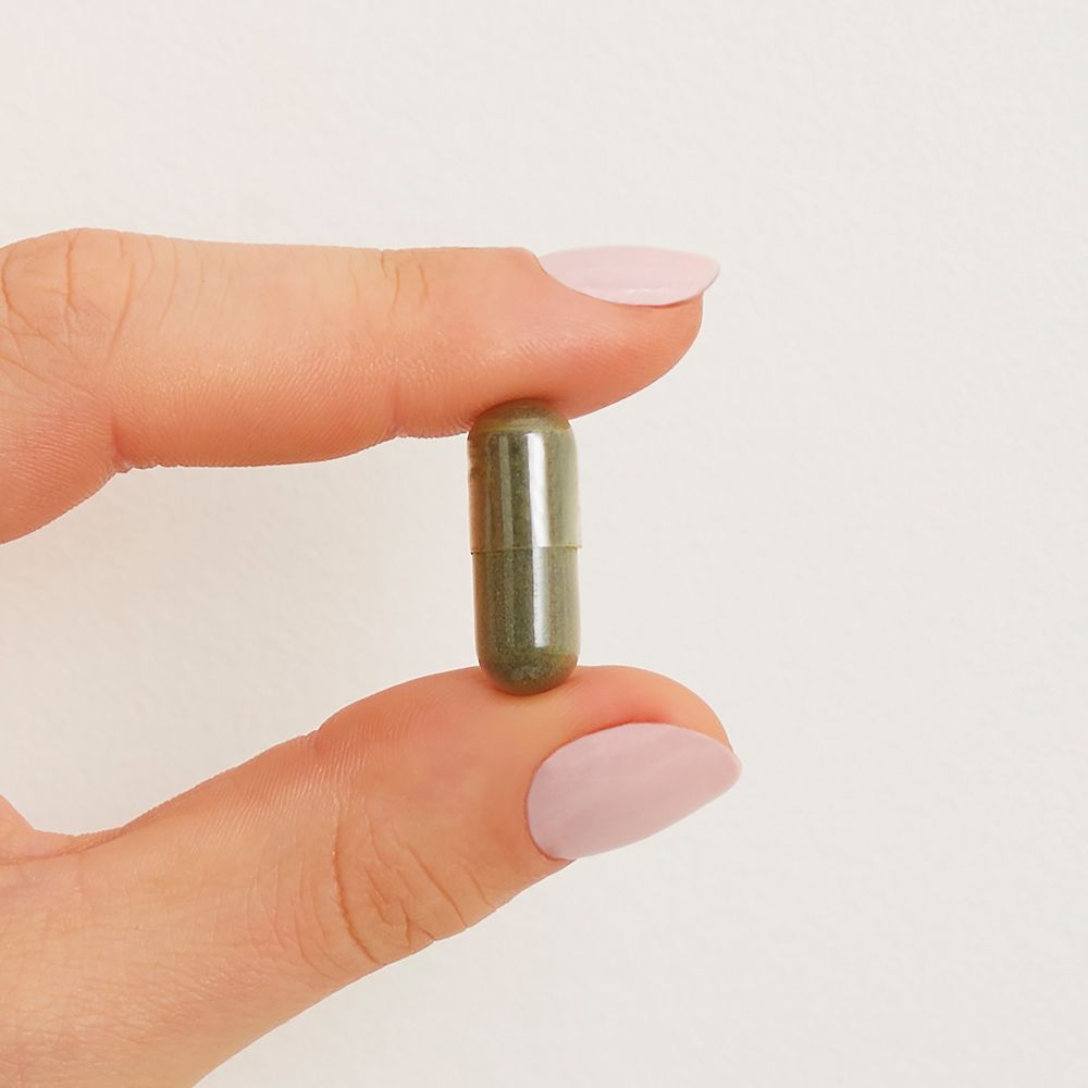 Main aux ongles roses tenant une gélule verte de Chlorella entre le pouce et l'index.