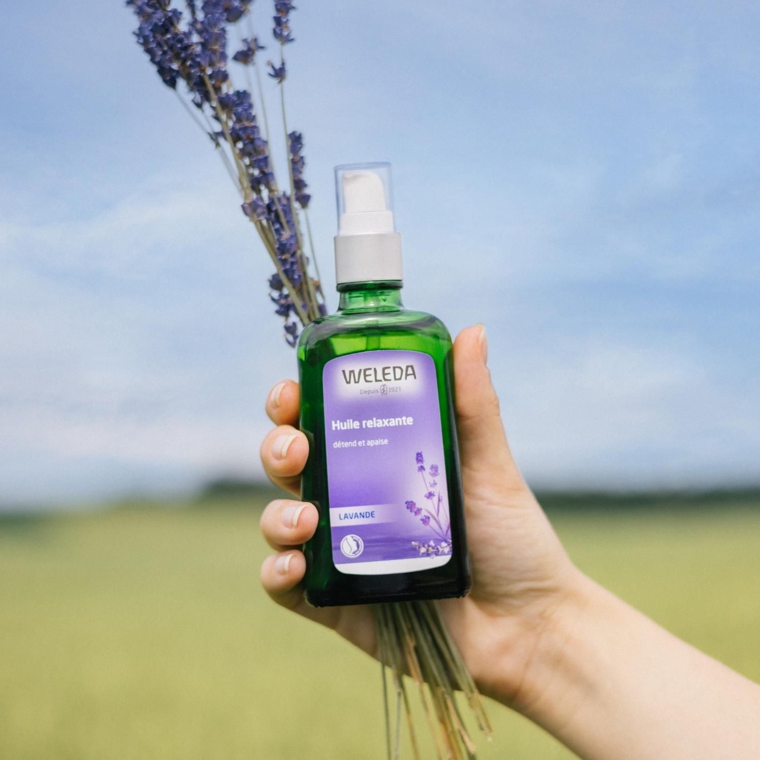 Hand hält Flasche Weleda Lavendel-Entspannungsöl und Lavendelblüten. Grüner Flakon mit Sprühkopf.