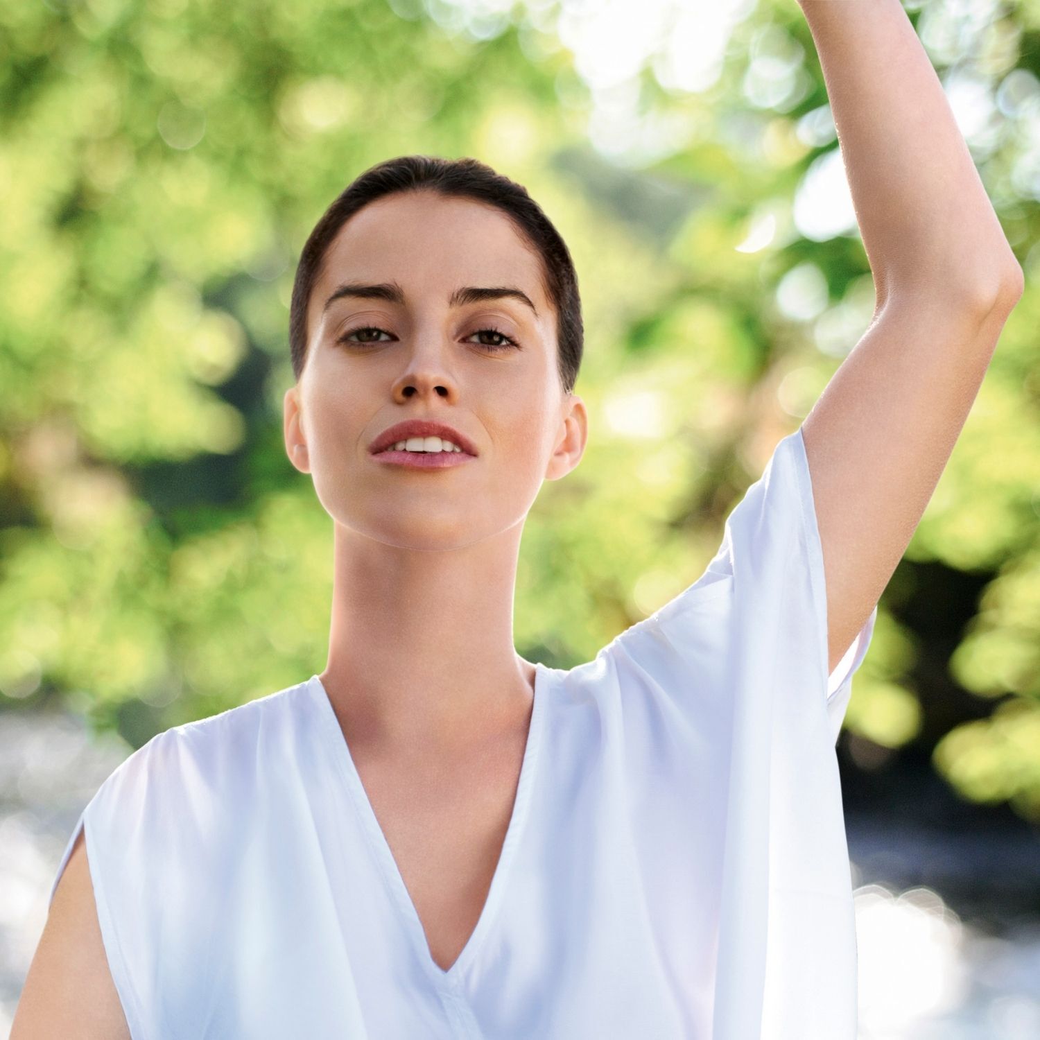 Eine Frau mit erhobenem Arm im Freien. Sie trägt ein weißes Kleid. Hintergrund: grünes Bokeh.
