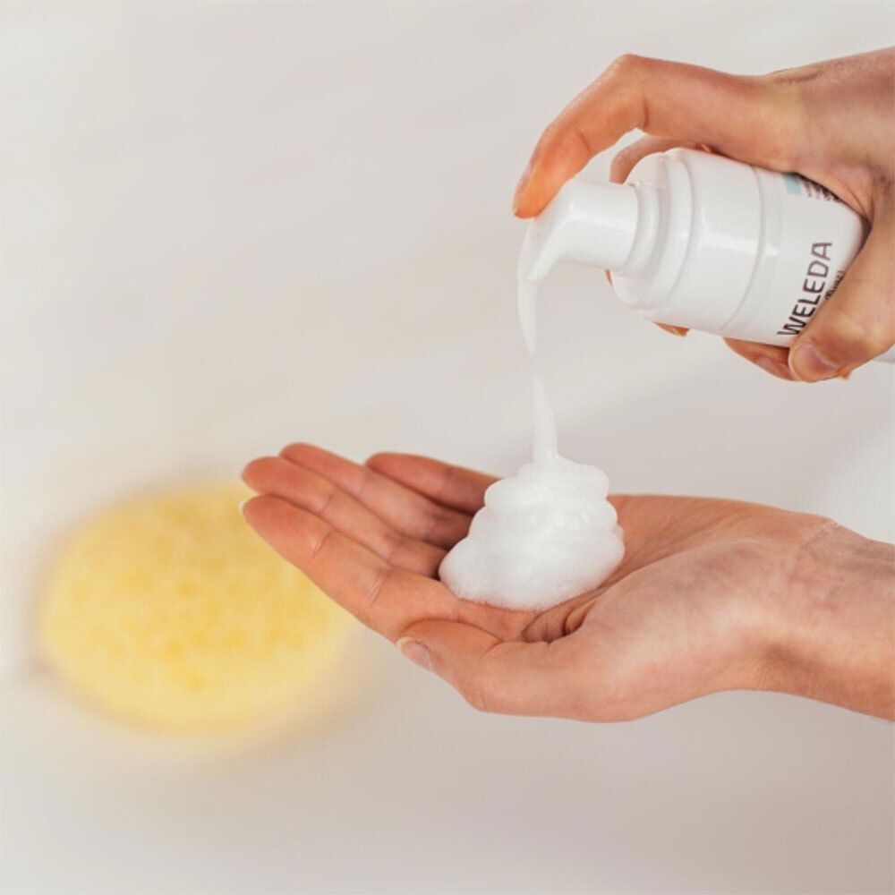 Mousse blanche sur la main. Éponge jaune en arrière-plan. Produit: WELEDA Gentle Cleansing Foam. Mousse sortant du flacon.