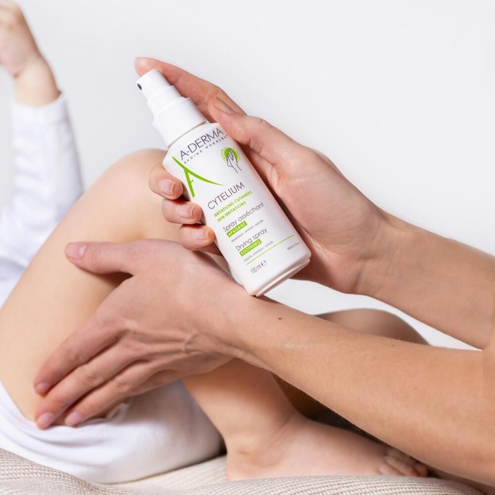 Mains tenant le spray asséchant A-DERMA CYTELIUM. Flacon pulvérisé sur une jambe. Flacon blanc avec étiquette verte.