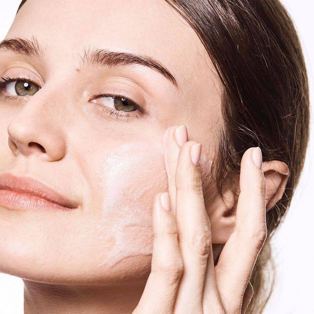 Gros plan sur un visage. Une main applique le produit sur la peau. Fond blanc.