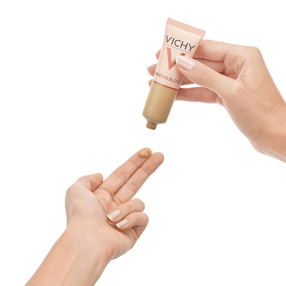 Main pressant le fond de teint VICHY Mineralblend Nr. 09 Achat. Produit sur le doigt. Tube rose-doré.