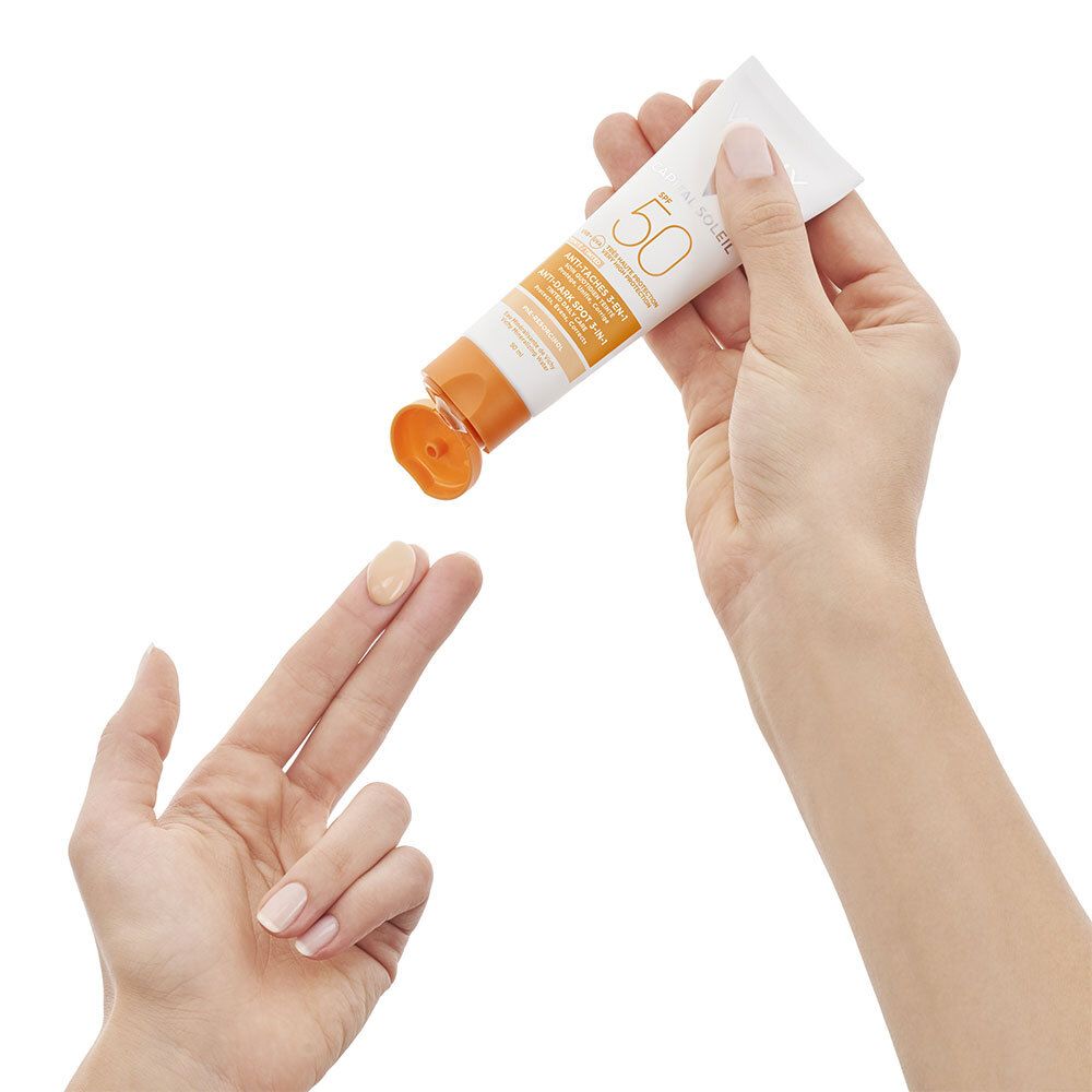 Hände halten eine Vichy Idéal Soleil 3-in-1 getönte Anti-Flecken-Pflege SPF 50+ Tube. Creme wird auf Finger aufgetragen.