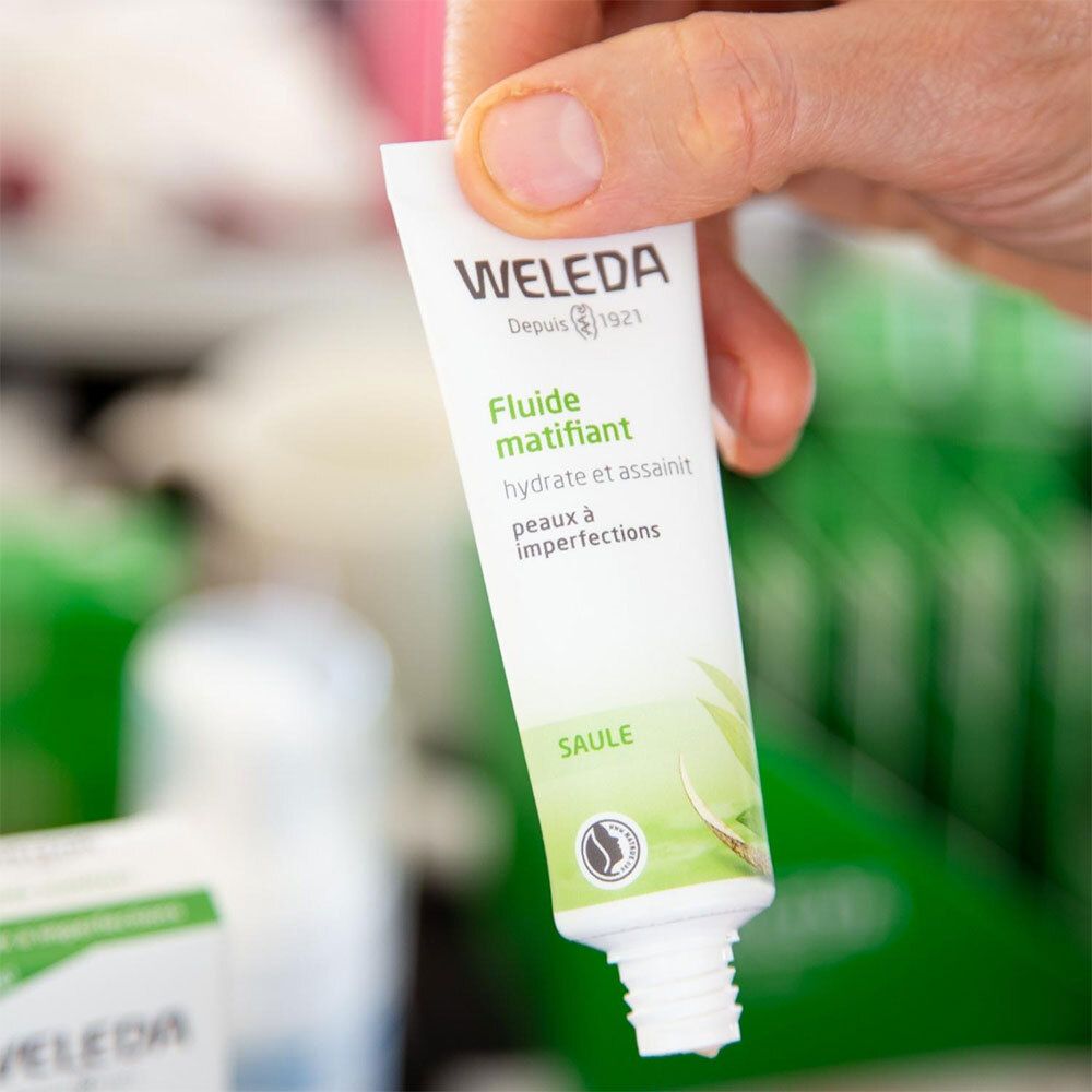 Main tenant un tube de fluide WELEDA. Impression : Fluiide matifiant. Avec logo et illustration végétale. Ouvert.