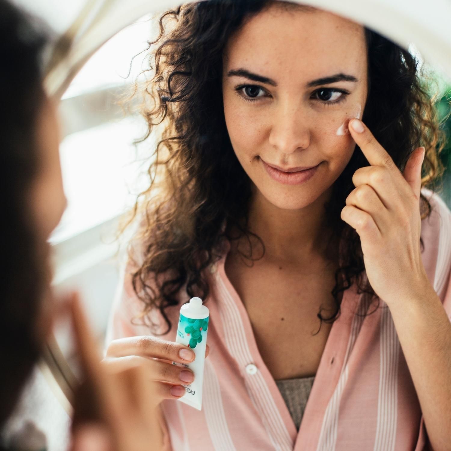 Femme applique crème. Tube vert. Reflet dans miroir. Produit partiellement visible.