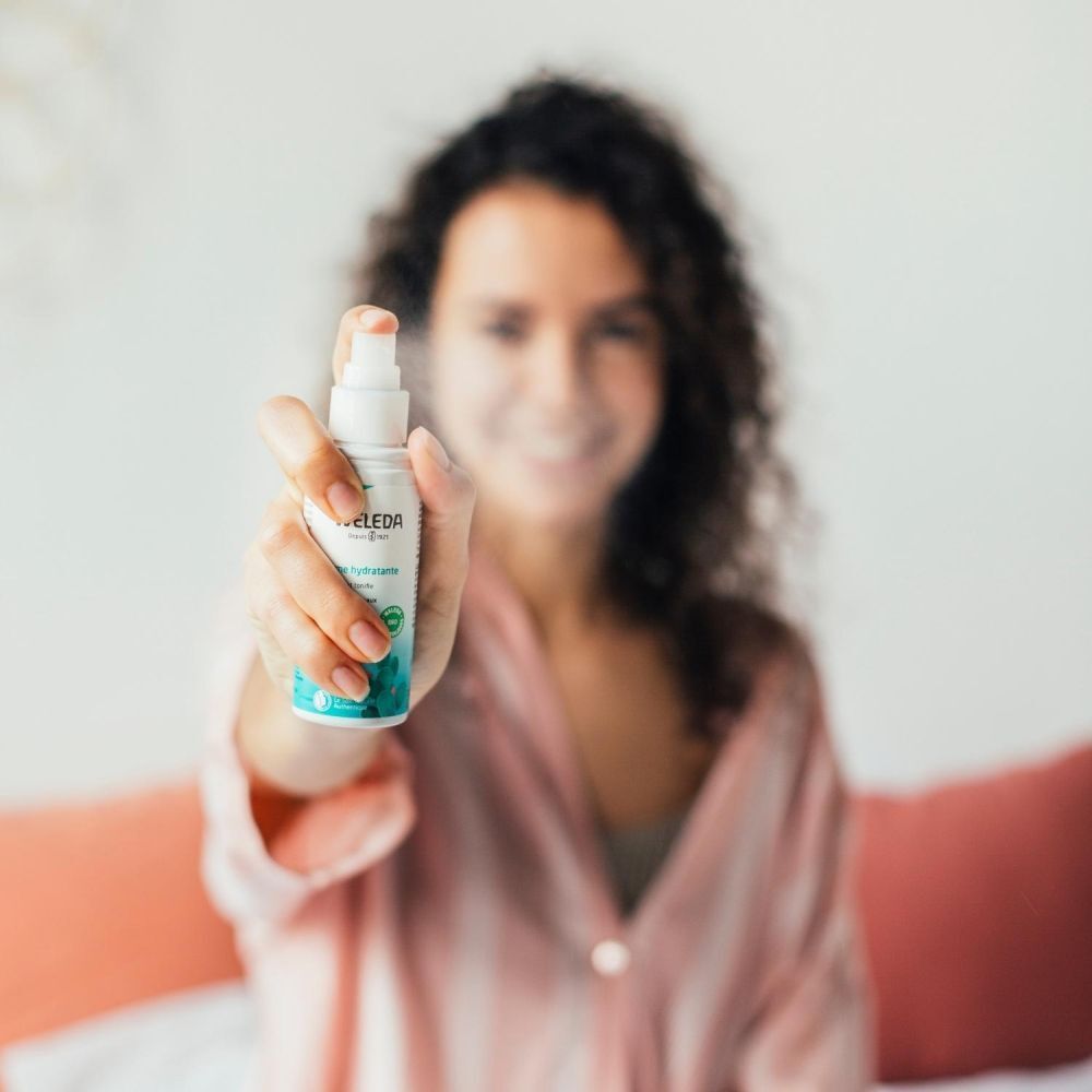 Femme tenant un flacon de spray Weleda vers la caméra. Le produit est flou, mais l'étiquette est visible.