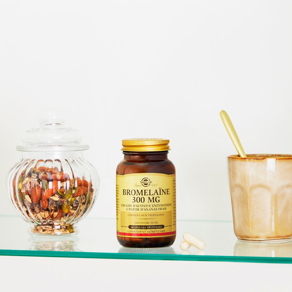 Scène avec un flacon de Bromelaïne, deux gélules et un verre sur une étagère. À côté d'un bocal et d'une tasse.